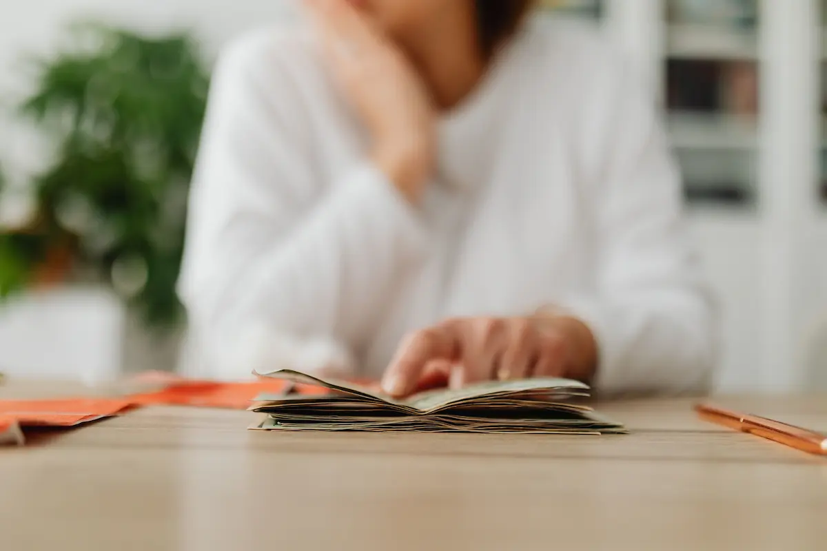Main posée sur une pile de billets près d'un stylo sur une table en bois, personne floue en arrière-plan